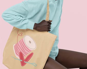 May include: A beige tote bag with a pink and white thread spool and a red heart design. The bag has two long handles.