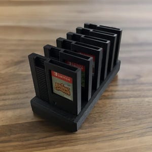 Puede incluir: Un soporte de plástico negro con ranuras para guardar cartuchos de juegos de Nintendo Switch. El soporte está diseñado para parecerse a una consola retro de Nintendo Entertainment System (NES). El soporte contiene varios cartuchos de juegos, incluido uno con la carátula del juego Animal Crossing: New Horizons.