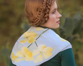 May include: A woman with long, red hair braided into a crown around her head wears a light blue scarf with a yellow floral print. The scarf is tied around her neck and drapes over her shoulders.