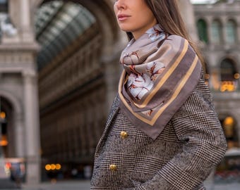 May include: A woman wearing a gray and white plaid blazer with gold buttons and a silk scarf. The scarf is taupe with a floral pattern and a gold border. The blazer is paired with tan pants.