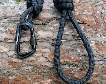 May include: Black rope with a black carabiner attached. The rope is tied in a knot and the carabiner is clipped to the rope. The rope and carabiner are lying on a piece of tree bark.