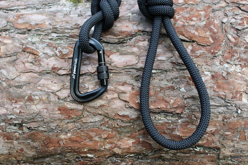 May include: Black rope with a black carabiner attached. The rope is tied in a knot and the carabiner is clipped to the rope. The rope and carabiner are lying on a piece of tree bark.