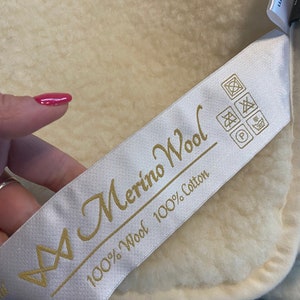 Könnte beinhalten: Weißes Etikett mit goldener Aufschrift "Merino Wool 100% Wool 100% Cotton". Das Etikett hat ein Symbol aus zwei Dreiecken und ein Waschsymbol.