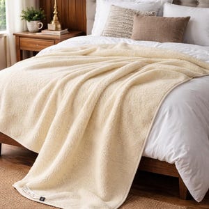 May include: A cream-colored, plush blanket draped over a bed. The blanket has a soft, textured appearance and is made of a thick, warm material. The bed has white linens and decorative pillows. The room has a warm, inviting atmosphere.