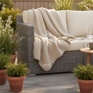May include: A beige, plush blanket draped over a gray wicker outdoor sofa. The blanket has a soft, textured appearance and a small label on the corner. Potted plants and a small side table with a mug are also visible.