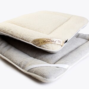 Könnte beinhalten: Ein weißer Merinowolle-Matratzenauflage mit einer grauen Baumwollunterseite. Die Auflage hat eine weiße Einfassung und weiße Bänder. Das Etikett lautet "Merino Wool 100% Natural Wool Fiber"