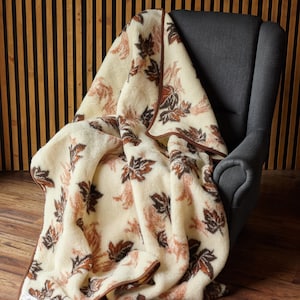 May include: A cream-colored merino wool blanket draped over a gray armchair. The blanket features a repeating pattern of brown and rust-colored leaves. The blanket has a brown border and a tag in the corner.