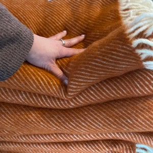 May include: A folded rust-orange throw blanket featuring a diagonal white herringbone pattern and cream-colored fringe at the edges. A small white rectangular fabric label is stitched onto the top corner of the textured, woven textile material.