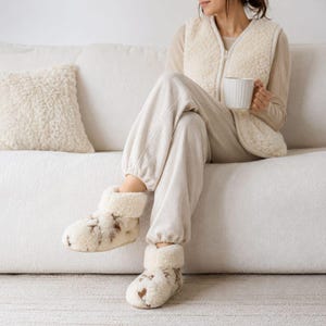 Könnte beinhalten: Gemütliche cremefarbene Fleece-Hausschuhe mit braunen Flecken. Die Hausschuhe haben einen umgeschlagenen Saum und werden mit einer passenden cremefarbenen Jogginghose und einer Fleece-Weste getragen. Eine weiße Tasse wird in der Hand gehalten.