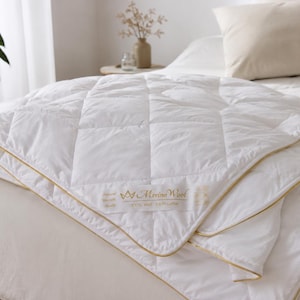 May include: A white, quilted comforter with gold trim is neatly folded on a bed. The comforter has a square pattern and the words "Merino Wool" are visible on a label. Two beige pillows rest on the bed, and a potted plant is in the background.
