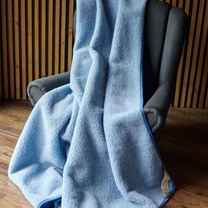 May include: A light blue merino wool blanket draped over a gray armchair. The blanket has a soft, fluffy texture with a contrasting blue trim. The blanket is on a wooden floor with a vertical wood panel background.