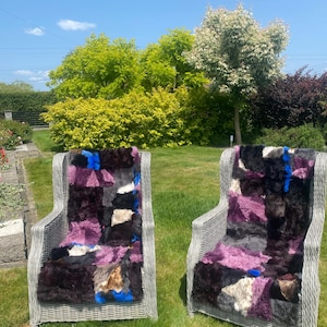 Op de afbeelding: Twee grijze rotan fauteuils met patchwork bontworpen. De worpen zijn gemaakt van paarse, zwarte, blauwe en bruine stukken bont. De stoelen staan op een groen gazon, met bomen en een blauwe lucht op de achtergrond, wat een gezellige buitensetting creëert.