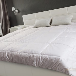 May include: A white bed with a white duvet cover and two white pillows. The bed is in a bedroom with a white crib in the background.