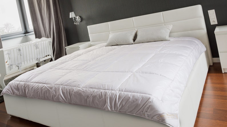 May include: A white bed with a white duvet cover and two white pillows. The bed is in a bedroom with a white crib in the background.