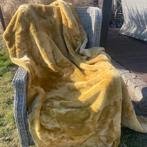 May include: A mustard yellow faux fur throw blanket with a visible patchwork square pattern. The blanket has a soft, plush texture and is draped over a light gray woven outdoor chair, showing deep folds and shadows across its surface.