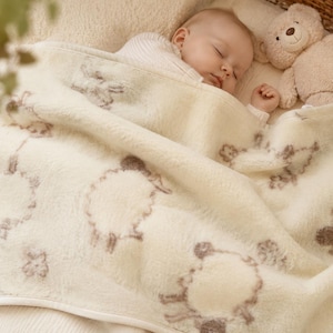 May include: A sleeping baby wrapped in a cream-colored blanket with a sheep pattern. A beige teddy bear rests beside the baby. The blanket has a tag that says "Merino Wool".