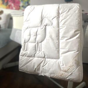 Puede incluir: Manta de bebé acolchada blanca con un diseño de oveja y el texto "Merino Wool 100% Wool 100% Cotton" en letras doradas.