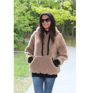 Puede incluir: Una sudadera con capucha de gran tamaño de color beige con un bolsillo de canguro y cordones negros. La sudadera tiene un aspecto suave y texturizado y se combina con una camiseta interior negra y vaqueros azules. La persona lleva gafas de sol.