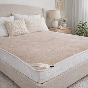 Könnte beinhalten: Eine beige, texturierte Matratzenauflage mit weißer Stoffkante und einem kleinen weißen Etikett an der Ecke, die fest auf einer weißen Matratze liegt. Die Oberfläche hat ein dichtes, vliesartiges Aussehen mit einem gleichmäßigen Flor.