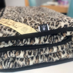 Może przedstawiać: Złożony koc z wzorem w cętki w odcieniach beżu, brązu i czerni. Koc ma czarną obwódkę i metkę z napisem "Merino Wool".