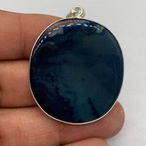 May include: A round pendant with a silver-colored border. The pendant's face features a dark blue and teal swirling pattern. The pendant is held by a hand, showcasing its size and design. The pendant has a small silver loop at the top for a chain.