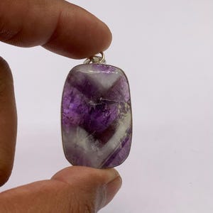 May include: A rectangular pendant featuring a polished amethyst stone with distinct bands of deep purple and opaque white. The gemstone is encased in a simple silver-toned metal frame with a small circular bail at the top for attachment to a chain.