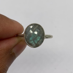 May include: A silver ring featuring an oval-shaped, faceted labradorite gemstone. The stone displays a grey base color with iridescent blue-green flashes visible across its surface. The metal band is thin, smooth, and polished in a metallic silver tone.