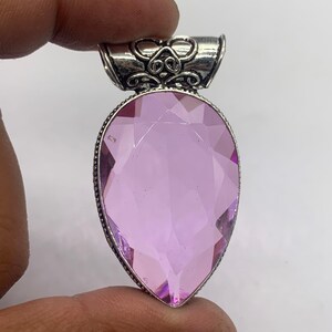 Può includere: Un ciondolo con pietra preziosa rosa chiaro sfaccettata a forma di pera, incastonato in una montatura in metallo color argento. La contromaglia presenta complessi motivi a volute e il bordo della pietra ha una texture perlata dettagliata.