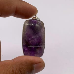 May include: A rectangular pendant featuring a polished purple amethyst gemstone with natural white inclusions and a horizontal vein, encased in a simple silver-toned metal frame with a small circular bail at the top for attachment to a chain.
