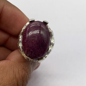 Puede incluir: Un anillo de plata con una gran piedra preciosa ovalada de color rojo oscuro. La piedra preciosa está engastada en una banda de plata detallada con un diseño texturizado. El anillo se muestra sobre un fondo blanco liso, resaltando las características de la joya.