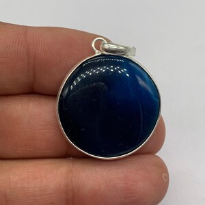 May include: A round, dark blue pendant with a silver-colored bail. The pendant has a polished, glossy finish and is set in a silver-colored metal frame. The blue color is a deep, rich shade.