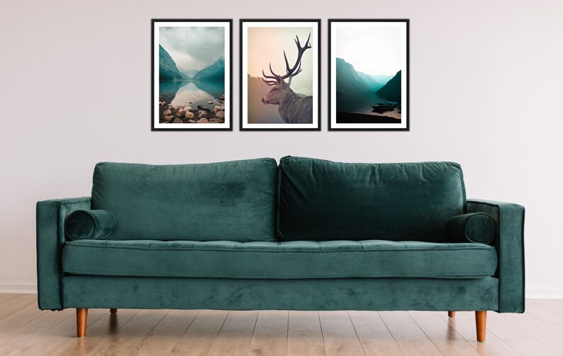May include: Three framed landscape photographs of a lake and mountains, a deer, and a lake and mountains. The photographs are hanging on a white wall above a teal green velvet sofa.