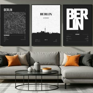 Peut inclure: Trois impressions d'art encadrées de Berlin. Les impressions présentent une carte, une silhouette d'horizon et du texte en gras, le tout en noir et blanc. Les impressions sont affichées au-dessus d'un canapé sectionnel gris avec des coussins orange et beiges.