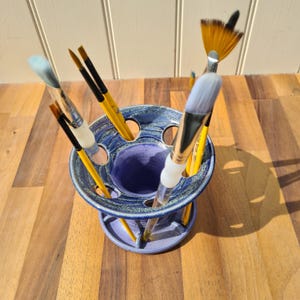 Peut inclure: Un porte-pinceaux en céramique bleu et violet, contenant plusieurs pinceaux. Les pinceaux ont des manches jaunes et des poils blancs ou beiges. Le support est posé sur une surface en bois, avec des ombres.