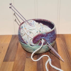 May include: A ceramic yarn bowl with white speckled yarn and two silver knitting needles. The bowl has a purple rim and a green base. Yarn exits the bowl through a curved opening.