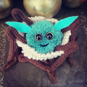 K&ouml;nnte beinhalten: Ein t&uuml;rkisfarbener Pompon mit gro&szlig;en schwarzen Augen und einem l&auml;chelnden Mund ist in einem braunen Stricknest eingebettet. Das Nest ist mit wei&szlig;em Garn verziert.