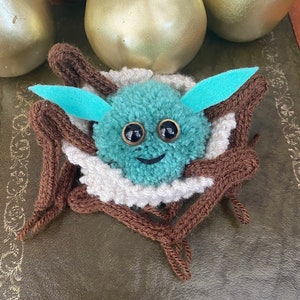 K&ouml;nnte beinhalten: Ein t&uuml;rkisfarbener Pompon mit Glitz Augen und einem l&auml;chelnden Mund ist in einem braunen und wei&szlig;en gestrickten Nest eingebettet. Das Nest ist aus Garn gefertigt und hat eine flauschige Textur.