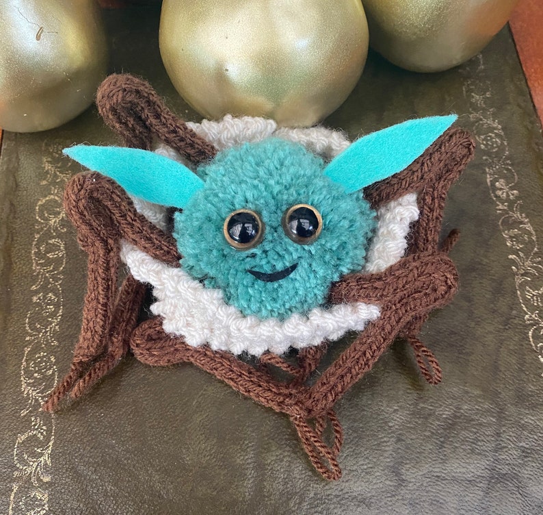 K&ouml;nnte beinhalten: Ein t&uuml;rkisfarbener Pompon mit Glitz Augen und einem l&auml;chelnden Mund ist in einem braunen und wei&szlig;en gestrickten Nest eingebettet. Das Nest ist aus Garn gefertigt und hat eine flauschige Textur.