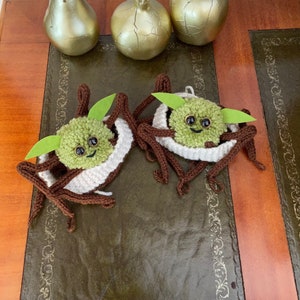 K&ouml;nnte beinhalten: Zwei geh&auml;kelte Yoda-Figuren in Gr&uuml;n und Braun mit wei&szlig;en Strickm&uuml;tzen auf einem braunen Holztisch mit goldenen Dekor-Birnen und einem Granatapfel.