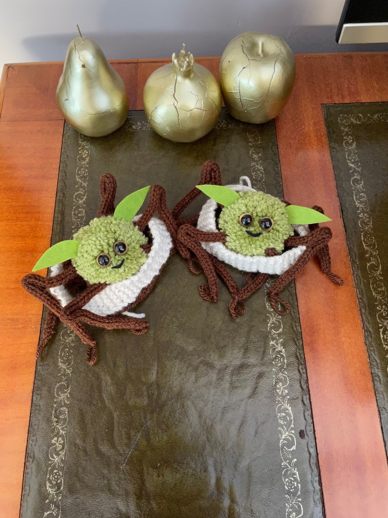 K&ouml;nnte beinhalten: Zwei geh&auml;kelte Yoda-Figuren in Gr&uuml;n und Braun mit wei&szlig;en Strickm&uuml;tzen auf einem braunen Holztisch mit goldenen Dekor-Birnen und einem Granatapfel.