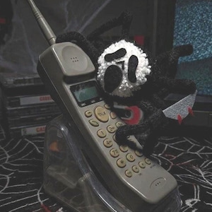 May include: A grey cordless telephone with a keypad and antenna sits in a clear plastic charging base. A black and white crocheted spider-like figure with a mask-like face and a small white knife with red accents is perched on the telephone.