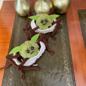 K&ouml;nnte beinhalten: Zwei geh&auml;kelte gr&uuml;n-braune Yoda-&auml;hnliche Figuren mit gro&szlig;en Augen und einem l&auml;chelnden Mund. Die Figuren sitzen auf einer braunen Lederoberfl&auml;che mit goldenen Dekorationselementen.