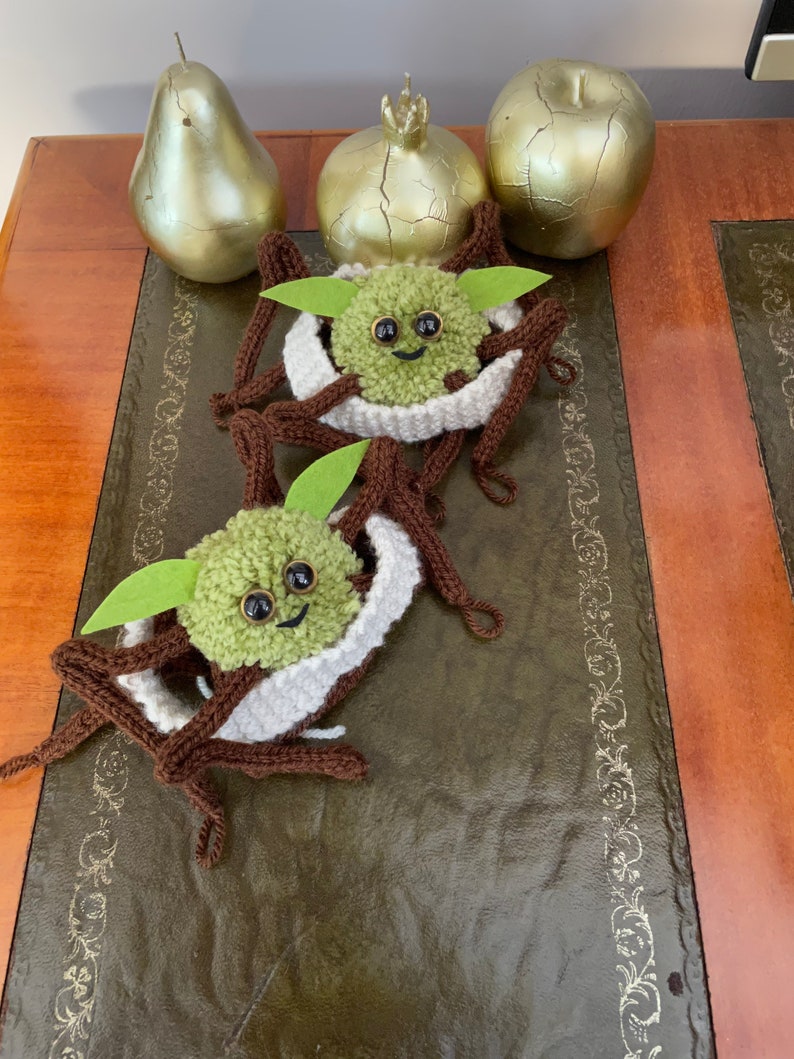 K&ouml;nnte beinhalten: Zwei geh&auml;kelte gr&uuml;n-braune Yoda-&auml;hnliche Figuren mit gro&szlig;en Augen und einem l&auml;chelnden Mund. Die Figuren sitzen auf einer braunen Lederoberfl&auml;che mit goldenen Dekorationselementen.