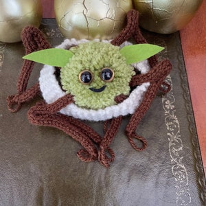 K&ouml;nnte beinhalten: Eine gr&uuml;ne gestrickte Garnfigur mit braunen Garnbeinen und einem wei&szlig;en Garnnest. Die Figur hat gro&szlig;e schwarze Augen und einen l&auml;chelnden Mund. Die Figur sitzt auf einer braunen Lederoberfl&auml;che.