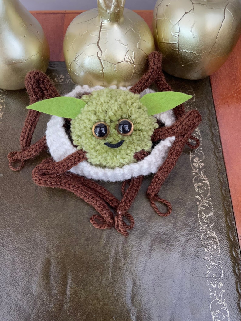K&ouml;nnte beinhalten: Eine gr&uuml;ne gestrickte Garnfigur mit braunen Garnbeinen und einem wei&szlig;en Garnnest. Die Figur hat gro&szlig;e schwarze Augen und einen l&auml;chelnden Mund. Die Figur sitzt auf einer braunen Lederoberfl&auml;che.