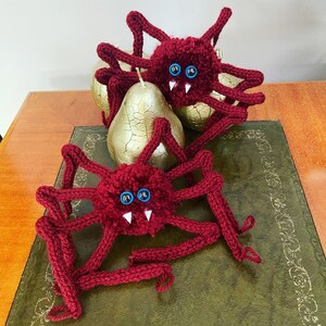 May include: Two red knitted spiders with googly eyes and fangs. The spiders are sitting on a green surface with a gold pear in the background.