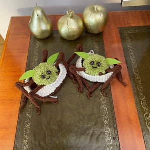 K&ouml;nnte beinhalten: Zwei geh&auml;kelte gr&uuml;n-braune Yoda-&auml;hnliche Figuren mit wei&szlig;en Garn-Akzenten. Die Figuren sitzen auf einem braunen Holztisch mit einer gr&uuml;nen Lederoberfl&auml;che.