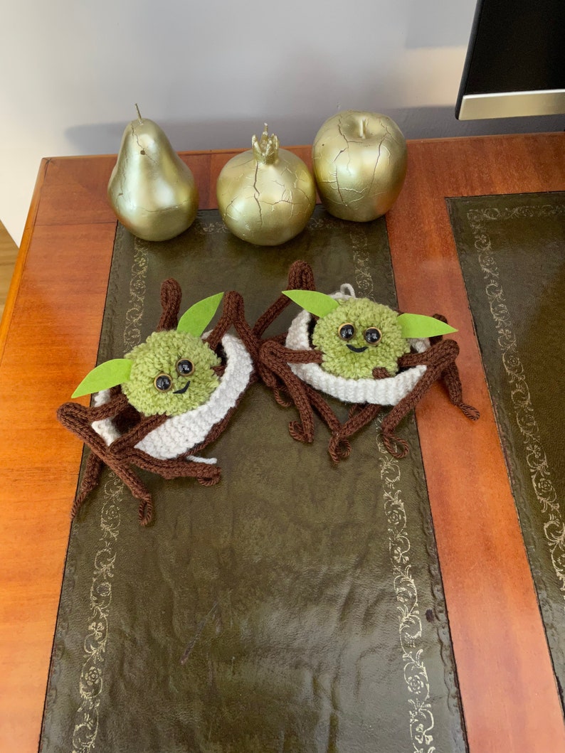 K&ouml;nnte beinhalten: Zwei geh&auml;kelte gr&uuml;n-braune Yoda-&auml;hnliche Figuren mit wei&szlig;en Garn-Akzenten. Die Figuren sitzen auf einem braunen Holztisch mit einer gr&uuml;nen Lederoberfl&auml;che.