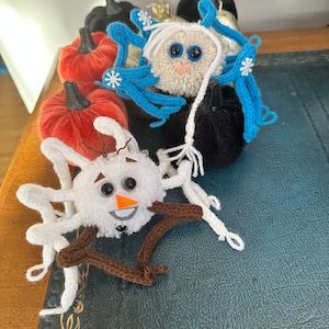 May include: A collection of handmade felt pumpkins decorated with blue and white yarn, snowflakes, and pom-poms. The pumpkins are arranged on a dark blue leather surface.