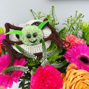 K&ouml;nnte beinhalten: Eine gr&uuml;ne geh&auml;kelte Spinne mit Glitz Augen und einem l&auml;chelnden Mund sitzt in einer Vase mit rosa, orangefarbenen und wei&szlig;en Blumen.